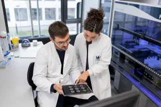 Researchers in a bright, glass-walled lab discuss cell therapy results over a digital tablet, with sophisticated lab devices operating in the background.