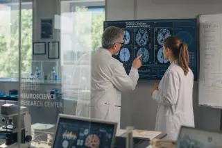 Two clinical researchers analyze digital brain scans and data on a screen in a modern medical research center, seen through a glass partition with soft daylight.